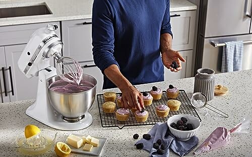 Person decorating cupcakes with frosting and blackberries in a kitchen